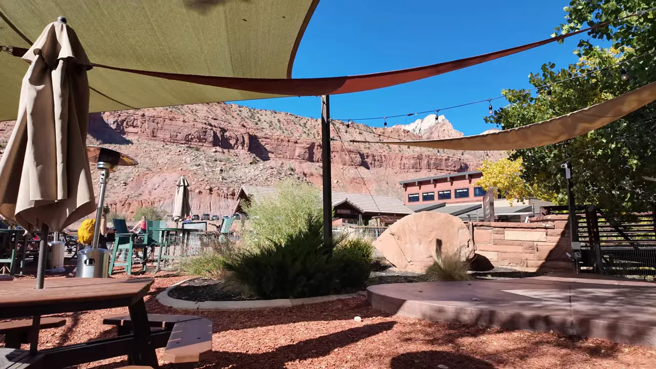 Empty outdoor brewery seating area showing shade sails, umbrellas and red sandstone cliffs beyond the patio.